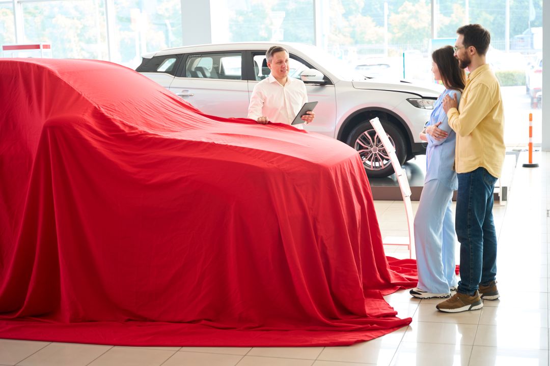 car dealership flags, a couple standing beside a featured car covered with a red blanket